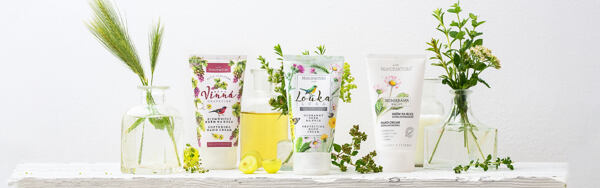 Tri krémy na ruky Manufaktura – Vínny, Lúka a Sedmokráska – postavené na bielej doske s vázami naplnenými zeleňou a kvetmi, doplnené o pohár s olejom a hrozna.