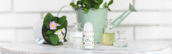 Prírodný roll-on dezodorant Manufaktura Lúka na stole s kanvičkou, kvetmi a sklenenými fľaštičkami.