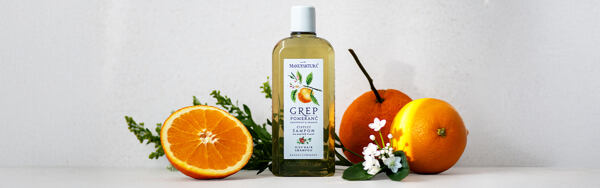 Fľaštička šampónu Manufaktura Grep a Pomaranč obklopená čerstvým grepom, pomarančom a kvetmi, na svetlom pozadí.