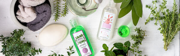 Dezinfekčné gély Manufaktura obklopené bylinkami, uterákmi a mydlom na svetlom drevenom podklade.