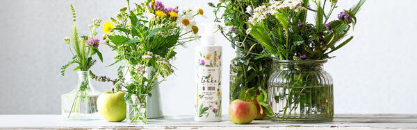 Telové mlieko Manufaktura Lúka obklopené jablkami, lúčnymi kvetmi a pohármi s bylinkami na drevenom podklade.