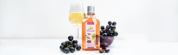 Fľaštička vínneho šampónu Manufaktura obklopená hroznovým vínom a pohárom bieleho vína na bielom pozadí.