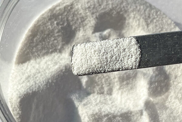 Biely prášok bambusového peelingu nabraný na čiernej plastovej špachtli nad miskou.