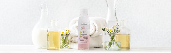 Dvoufázový odličovač Manufaktura se sedmikráskou, obklopený flakony s oleji a čerstvými květy na světlém pozadí.