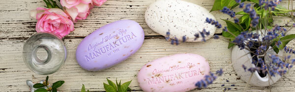 Tri tuhé mydlá Manufaktura vo fialovej, bielej a ružovej farbe, naaranžované s ružami, levanduľou a zeleňou na drevenom podklade.