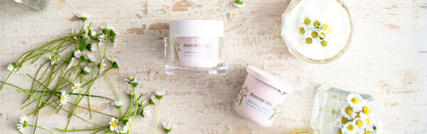 Denné a nočné pleťové krémy Manufaktura s motívom sedmokrásky, obklopené čerstvými kvietkami a nádobou s krémom.