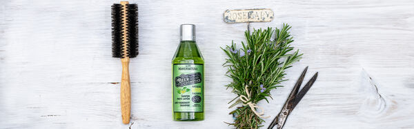 Fľaštička zeleného pivného šampónu Manufaktura proti lupinám, vedľa okrúhlej kefy, rozmarínu a nožníc na svetlom drevenom pozadí.