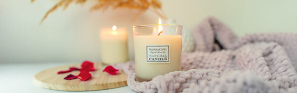 Horiaca prírodná sviečka Manufaktura v interiéri s pletenou dekou, okvetnými lístkami a tlmeným svetlom.