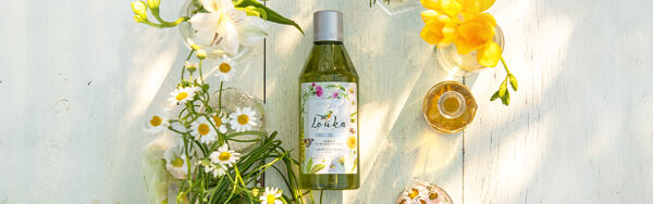 Sprchovací gél Manufaktura Lúka obklopený kvetmi, bylinkami a fľaštičkami s olejom na drevenom podklade.