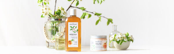 Šampón a maska Marhuľa Manufaktura uprostred kvetov a sklenených nádob, so zelenými vetvičkami v pozadí.