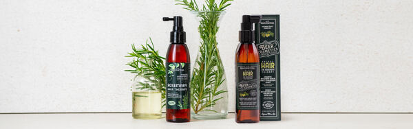 Dve fľaštičky vlasových sér Manufaktura s rozmarínom, doplnené vetvičkami bylín a sklenenými nádobami s olejom.