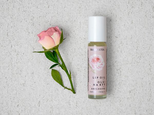 Ružový olej na pery od Manufaktury s ilustráciou kvetu ruže položený vedľa čerstvého ružového puku na svetlom povrchu