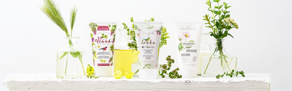 Tri krémy na ruky Manufaktura z kolekcií Vínna, Lúka a Sedmokráska, obklopené kvetmi a pohármi s olejmi.