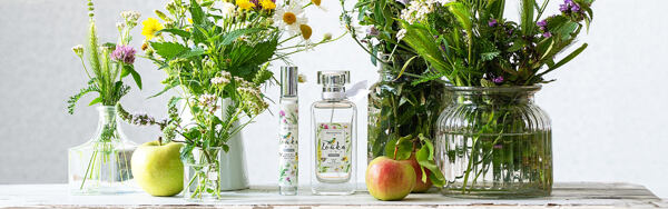 Dve dámske toaletné vody Lúka z Manufaktury medzi kvetinami v sklenených vázach a jablkami na drevenom stole.