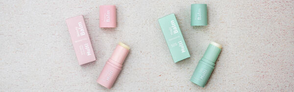 Dva balzamy na pery Manufaktura v tyčinke – ružový Multifunkčný balzam a zelený SOS balzam s príslušnými krabičkami na svetlom podklade.