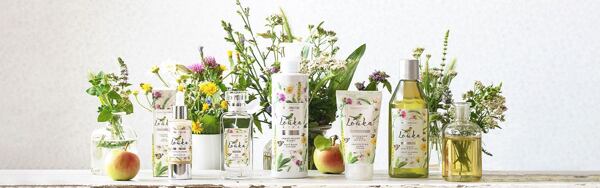 Kosmetické produkty Manufaktura z kolekce Louka obklopené lučními květinami, jablky a bylinkovými výtažky na světlém pozadí.