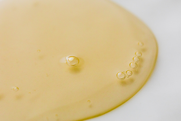 Detailný pohľad na zlatistú žltú tekutinu s niekoľkými malými bublinkami na hladkom bielom povrchu, pripomínajúcu ricínový olej.